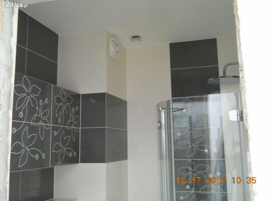 Wykończona łazienka z kabiną prysznicową z motywem kwiatowym, czarno-białe płytki z ornamentami kwiatowymi, wentylator na ścianie, widok z narożnika pomieszczenia.