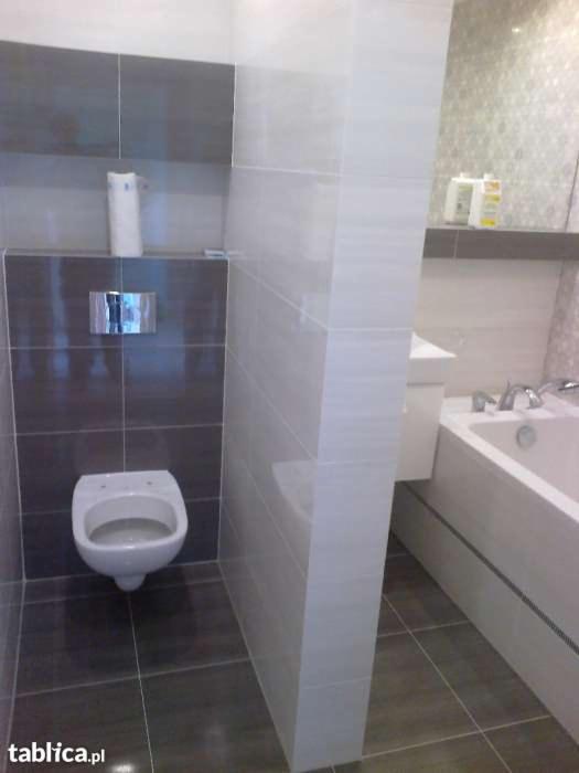 Nowoczesna łazienka z podwieszaną toaletą, szare i białe płytki, widoczna wanna z baterią i półka z kosmetykami.