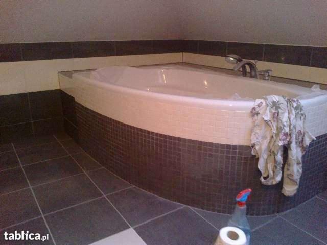 Wanna narożna z obudową z mozaiki w odcieniach brązu i beżu, na podłodze ciemne płytki, widoczny fragment ściany z pasem płytek, na wannie leży wzorzysty ręcznik, a na podłodze obok wanny stoi...