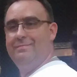 Portret mężczyzny w okularach, ubrany w białą koszulkę, na rozmytym tle.