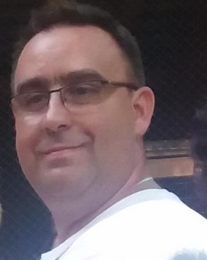 Portret mężczyzny w okularach, ubrany w białą koszulkę, na rozmytym tle.