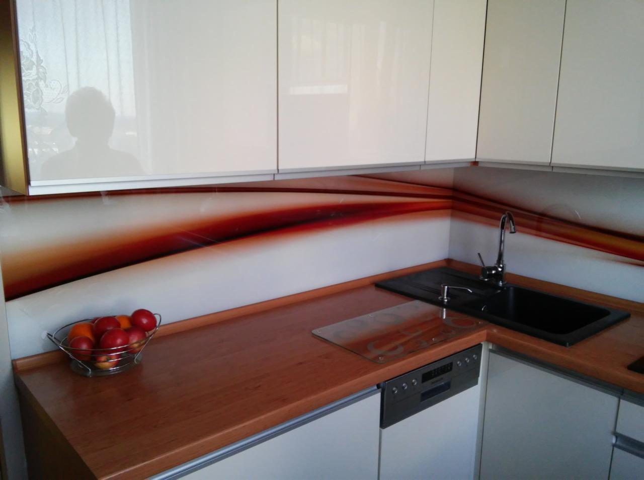 Białe szafki kuchenne z lakierowanymi frontami i szklanym panelem ściennym z abstrakcyjnym wzorem w odcieniach czerwieni i brązu, uzupełnione brązowym blatem i metalowym koszykiem z owocami.