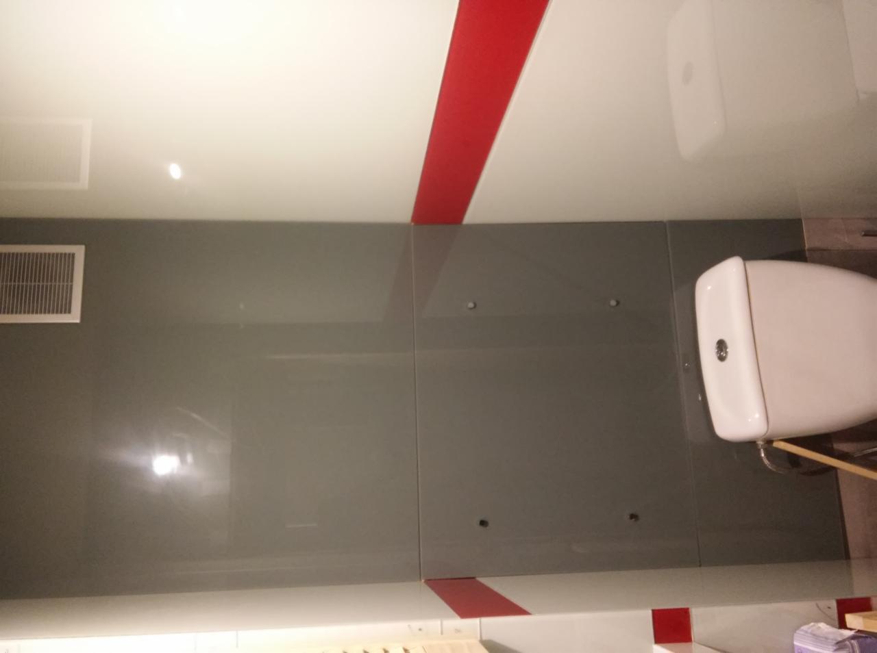 Szare szklane panele ścienne w łazience z czerwonymi akcentami i wiszącą toaletą, widoczne otwory montażowe w panelach.