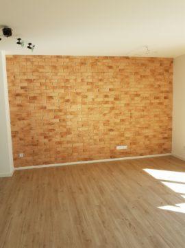 Pusta przestrzeń z nowo wykończoną ścianą imitującą cegłę i jasną podłogą z paneli, widoczne oświetlenie sufitowe.