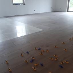 perfektee.pl - Układanie płytek podłogowych w jasnym, przestronnym wnętrzu, z widocznymi systemami poziomowania płytek w kolorze żółto-niebieskim, część podłogi już wyłożona jasnymi płytkami.