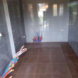 perfektee.pl - Szara łazienka w trakcie remontu z brązowymi płytkami na podłodze, widoczne wystające rury instalacji wodnej w kolorze niebiesko-czerwonym oraz ślady wilgoci na podłodze.