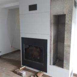 perfektee.pl - Nowoczesny kominek obudowany płytami imitującymi beton i dekoracyjnymi płytkami z ornamentem, widoczne cegły i poziomica użyte do montażu, podłoga wyłożona panelami.