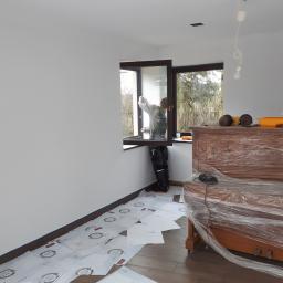 perfektee.pl - Pracownik w trakcie montażu okna w nowym, jasnym pomieszczeniu, zabezpieczone meble folią, podłoga wyłożona panelami i kartonami ochronnymi.