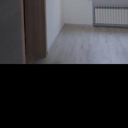 perfektee.pl - Jasne panele podłogowe w nowym, pustym pokoju z białymi ścianami, grzejnikiem pod oknem i fragmentem zabudowy szafy.