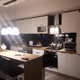 perfektee.pl - Nowoczesna kuchnia z białymi szafkami, czarną glazurą między blatem a szafkami wiszącymi, drewnianym blatem i wyspą z hokerami oraz oświetleniem wiszącym.