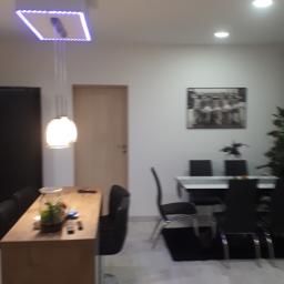 perfektee.pl - Nowoczesne wnętrze: drewniany blat z roślinami, oświetlenie LED i minimalistyczny stół z krzesłami na tle jasnej podłogi.