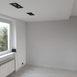 perfektee.pl - Pomalowane na biało puste pomieszczenie z panelami podłogowymi, grzejnikiem pod oknem i dwoma czarnymi reflektorami w suficie podwieszanym.