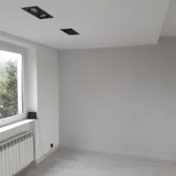 Pomalowane na biało puste pomieszczenie z panelami podłogowymi, grzejnikiem pod oknem i dwoma czarnymi reflektorami w suficie podwieszanym.