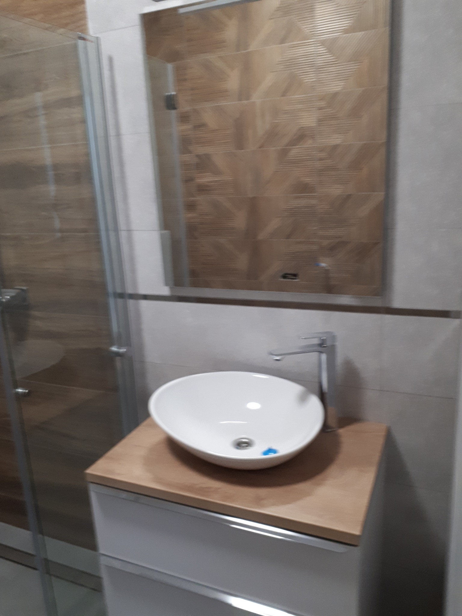 Nowoczesna łazienka z owalną umywalką nablatową, bateria chromowana, lustro z dekoracyjnymi płytkami imitującymi drewno i kabina prysznicowa z przesuwnymi drzwiami.