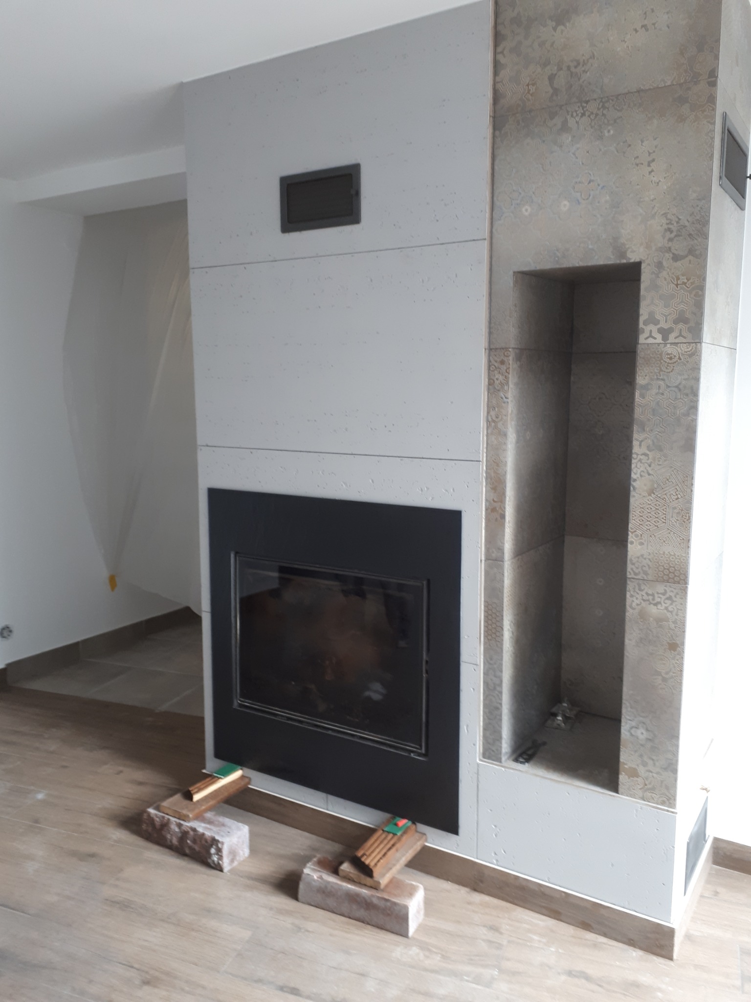 Nowoczesny kominek obudowany płytami imitującymi beton i dekoracyjnymi płytkami z ornamentem, widoczne cegły i poziomica użyte do montażu, podłoga wyłożona panelami.