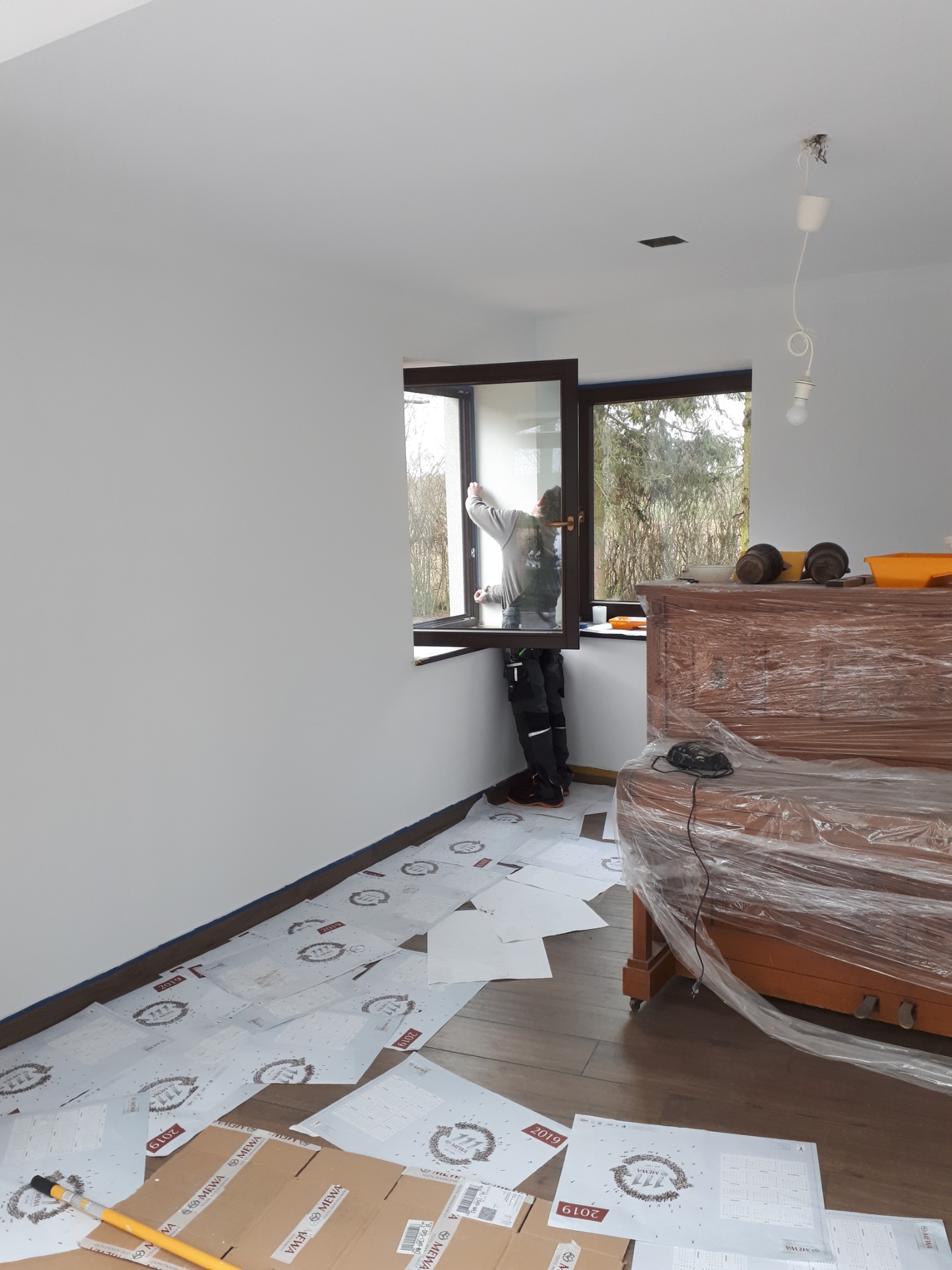 Pracownik w trakcie montażu okna w nowym, jasnym pomieszczeniu, zabezpieczone meble folią, podłoga wyłożona panelami i kartonami ochronnymi.