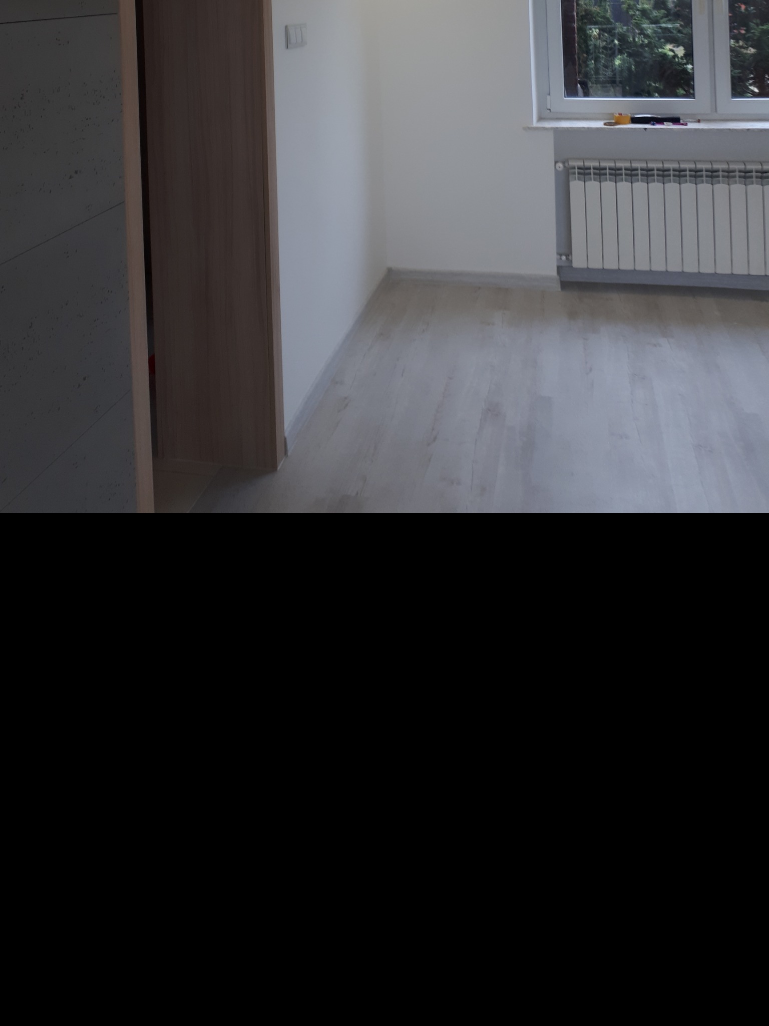 Jasne panele podłogowe w nowym, pustym pokoju z białymi ścianami, grzejnikiem pod oknem i fragmentem zabudowy szafy.