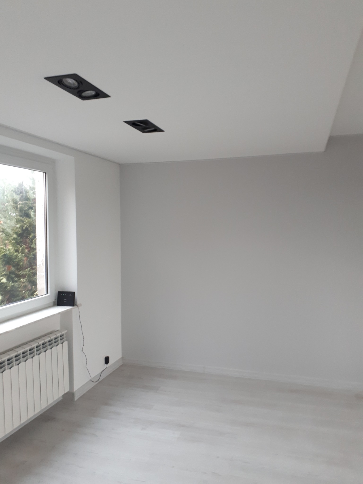 Pomalowane na biało puste pomieszczenie z panelami podłogowymi, grzejnikiem pod oknem i dwoma czarnymi reflektorami w suficie podwieszanym.