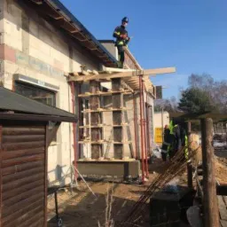 Wznoszenie ścian budynku z betonu komórkowego; widoczne szalunki i podpory, pracownik na dachu z dokumentacją, zbrojenie leżące na ziemi.