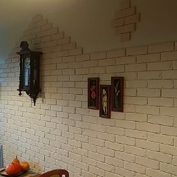 DmRem - Ściana wyłożona białymi cegłami dekoracyjnymi w salonie, z fragmentami cegieł tworzącymi dekoracyjny wzór na górze, wiszący zegar i trzy obrazy z motywami kwiatowymi.