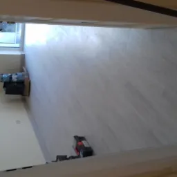 Jasne panele podłogowe ułożone w pustym pokoju, widoczne narzędzia do montażu i materiały budowlane przy ścianie