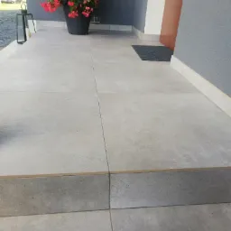 Jasnoszare płytki podłogowe na zewnątrz budynku, z donicą z czerwonymi kwiatami i czarnym lampionem dekoracyjnym