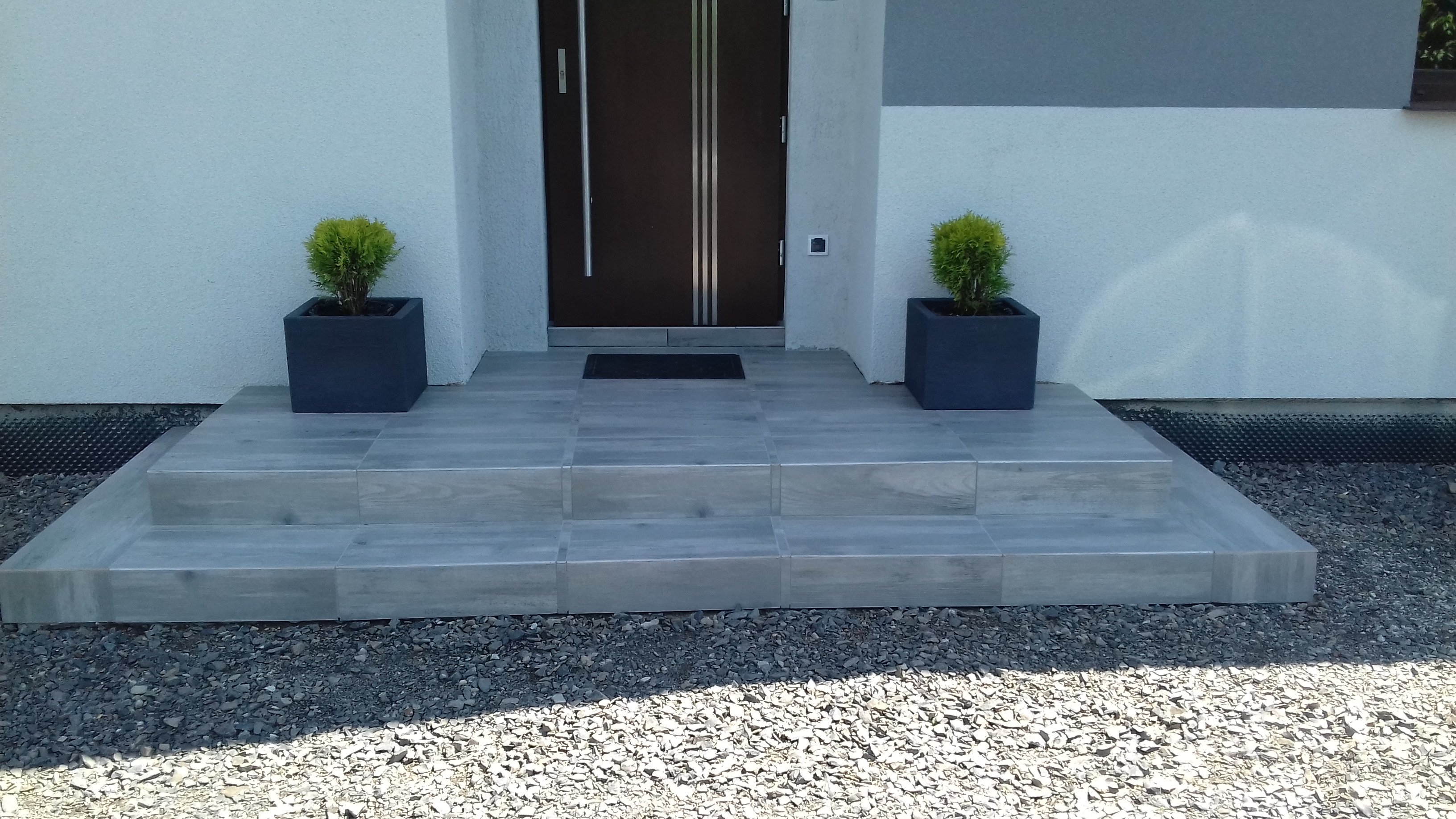 Wyłożone płytkami schody wejściowe do domu z ciemnymi drzwiami i dwoma donicami z tujami po bokach, na tle białej elewacji.