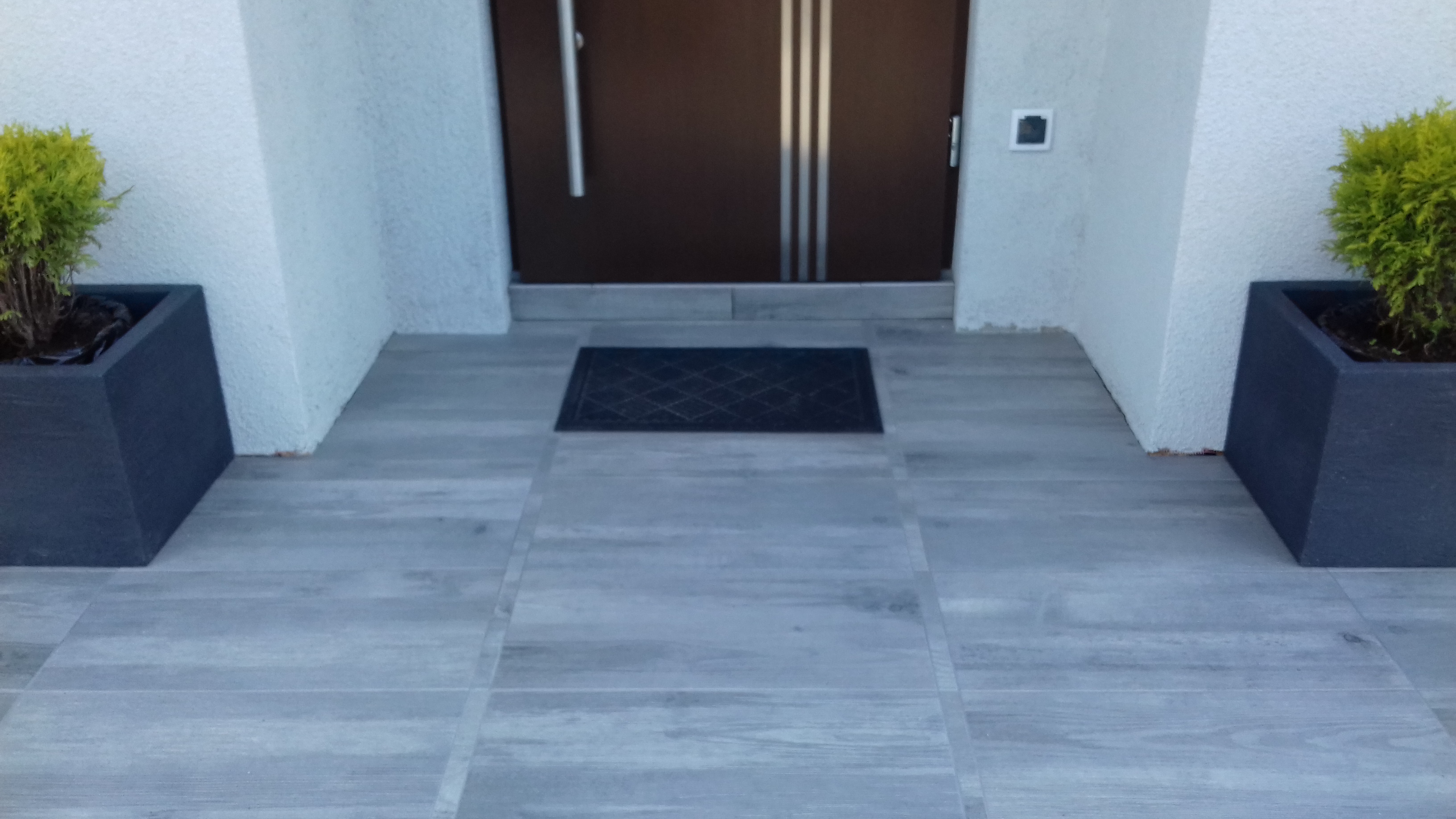 Szare, drewnopodobne płytki na zewnątrz prowadzące do brązowych drzwi wejściowych z metalowymi paskami, czarna wycieraczka i dwie czarne donice z zielonymi roślinami po bokach.