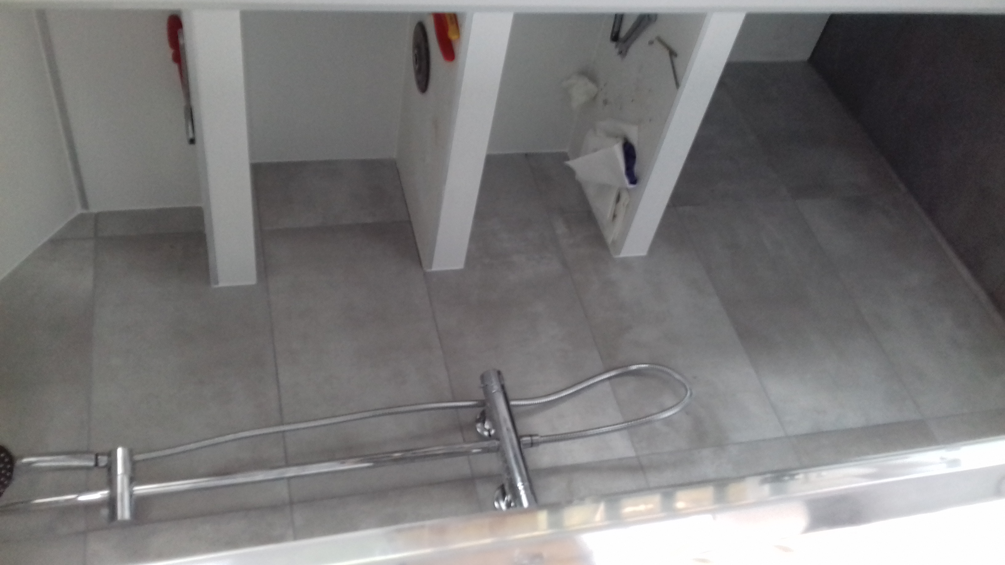 Szare płytki podłogowe w kabinie prysznicowej, widoczny chromowany panel prysznicowy i fragment ściany z białymi półkami i narzędziami.
