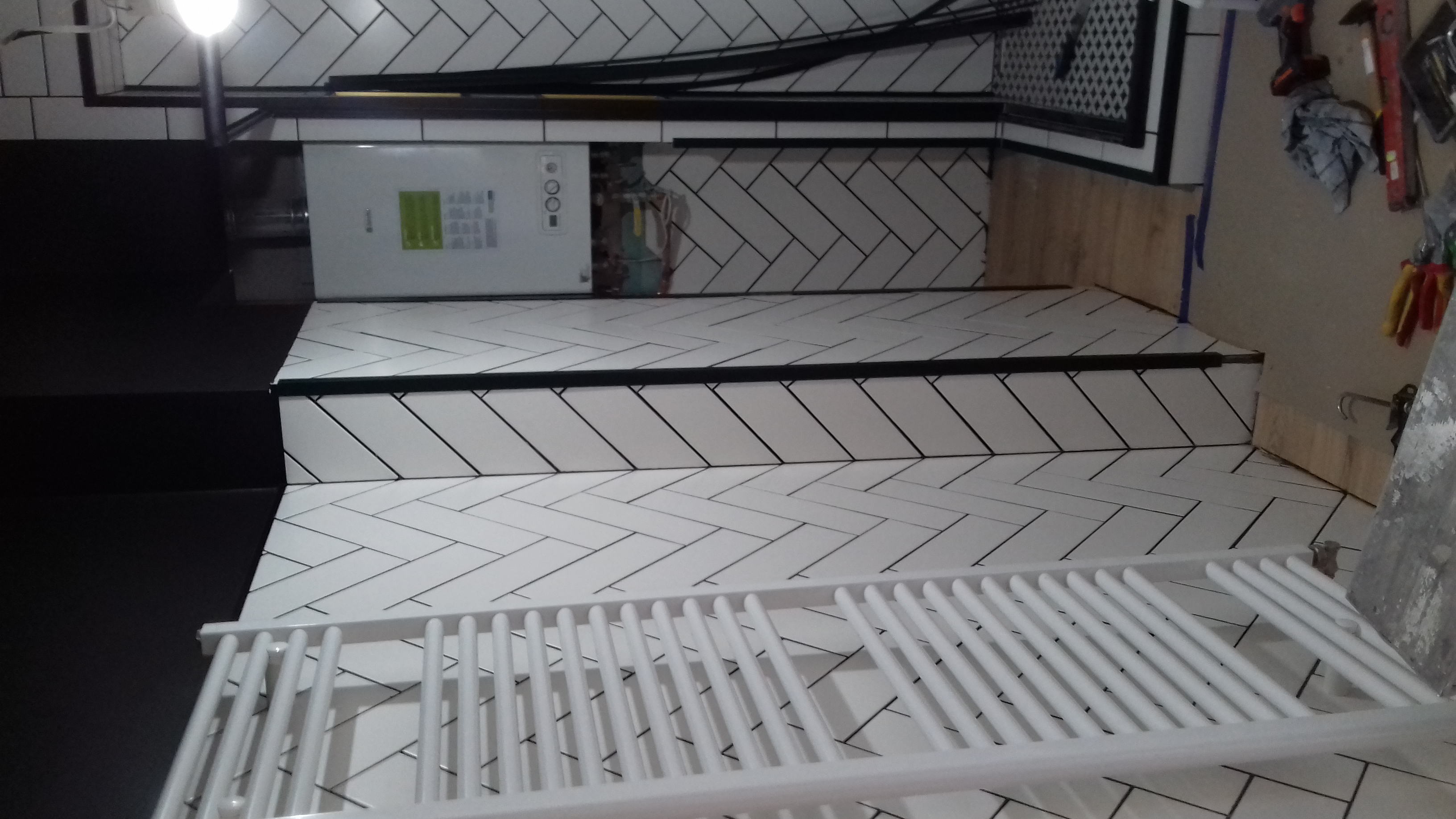 Białe, ułożone w jodełkę płytki ceramiczne na ścianie łazienki, z widocznym grzejnikiem drabinkowym i piecem gazowym oraz narzędziami w tle.