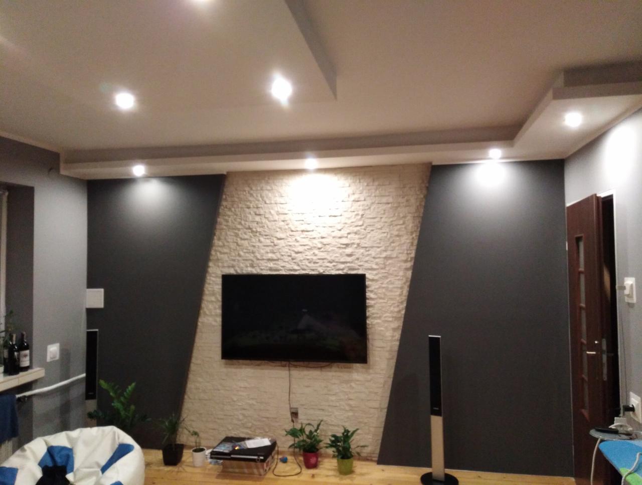 Salon z podświetloną ścianą z dekoracyjnej cegły, telewizorem i ciemnymi ścianami bocznymi, oświetlenie punktowe w suficie, rośliny doniczkowe na podłodze.