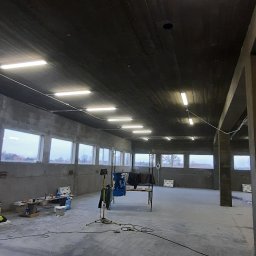 Elektro Instal Kamil Garbuliński - Wnętrze surowego, betonowego pomieszczenia z oświetleniem jarzeniowym; widoczne okna, narzędzia i prowizoryczne stanowisko pracy