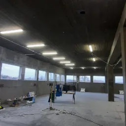 Wnętrze surowego, betonowego pomieszczenia z oświetleniem jarzeniowym; widoczne okna, narzędzia i prowizoryczne stanowisko pracy