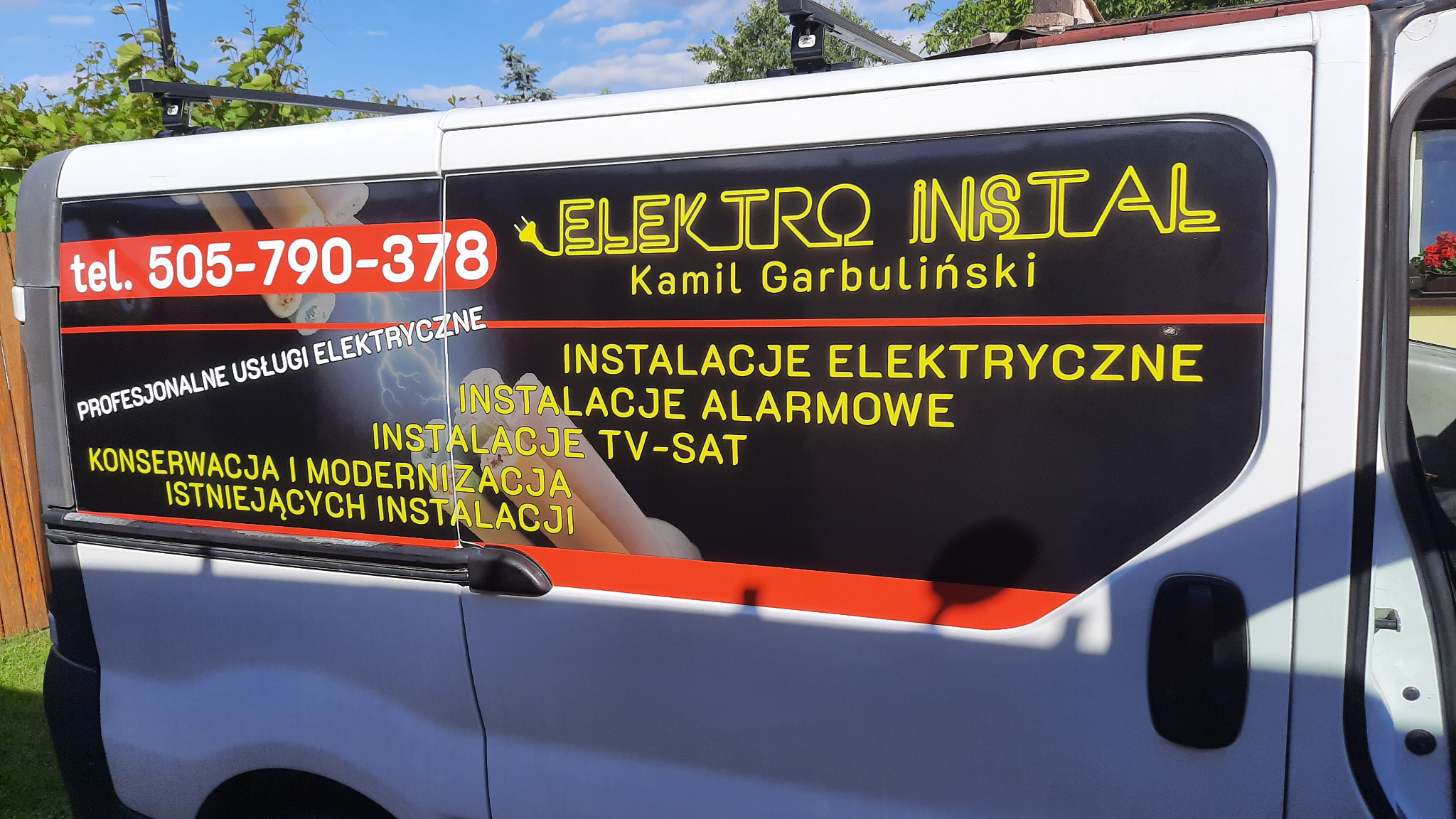 Biały samochód dostawczy z reklamą firmy Elektro Instal Kamil Garbuliński, oferującej profesjonalne usługi elektryczne, instalacje alarmowe i TV-SAT, konserwację i modernizację istniejących...