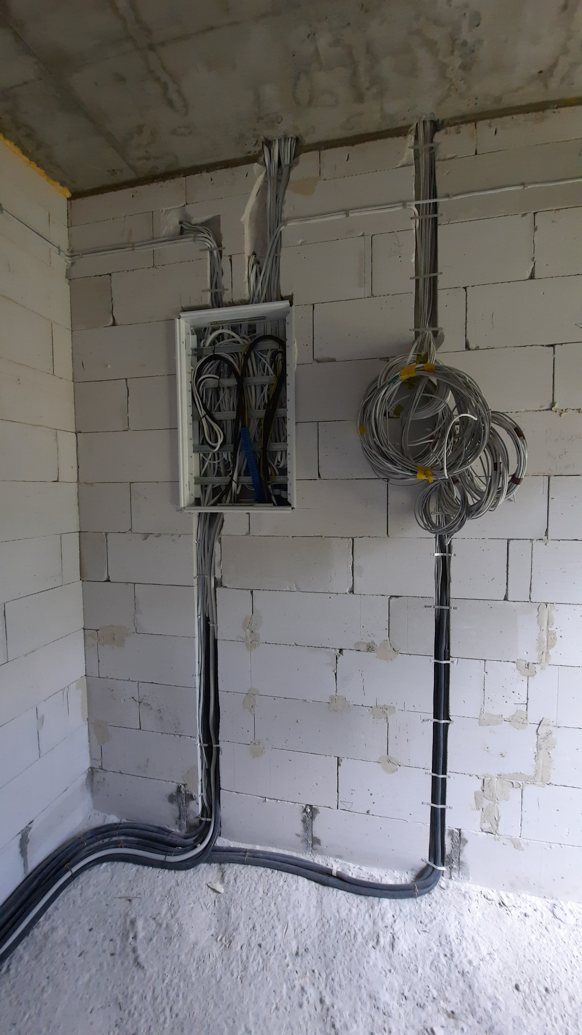 Rozdzielnica elektryczna z widocznymi przewodami w ścianie z białego bloczka, część przewodów zwinięta w pętle i przymocowana do ściany. Podłoga w stanie surowym.