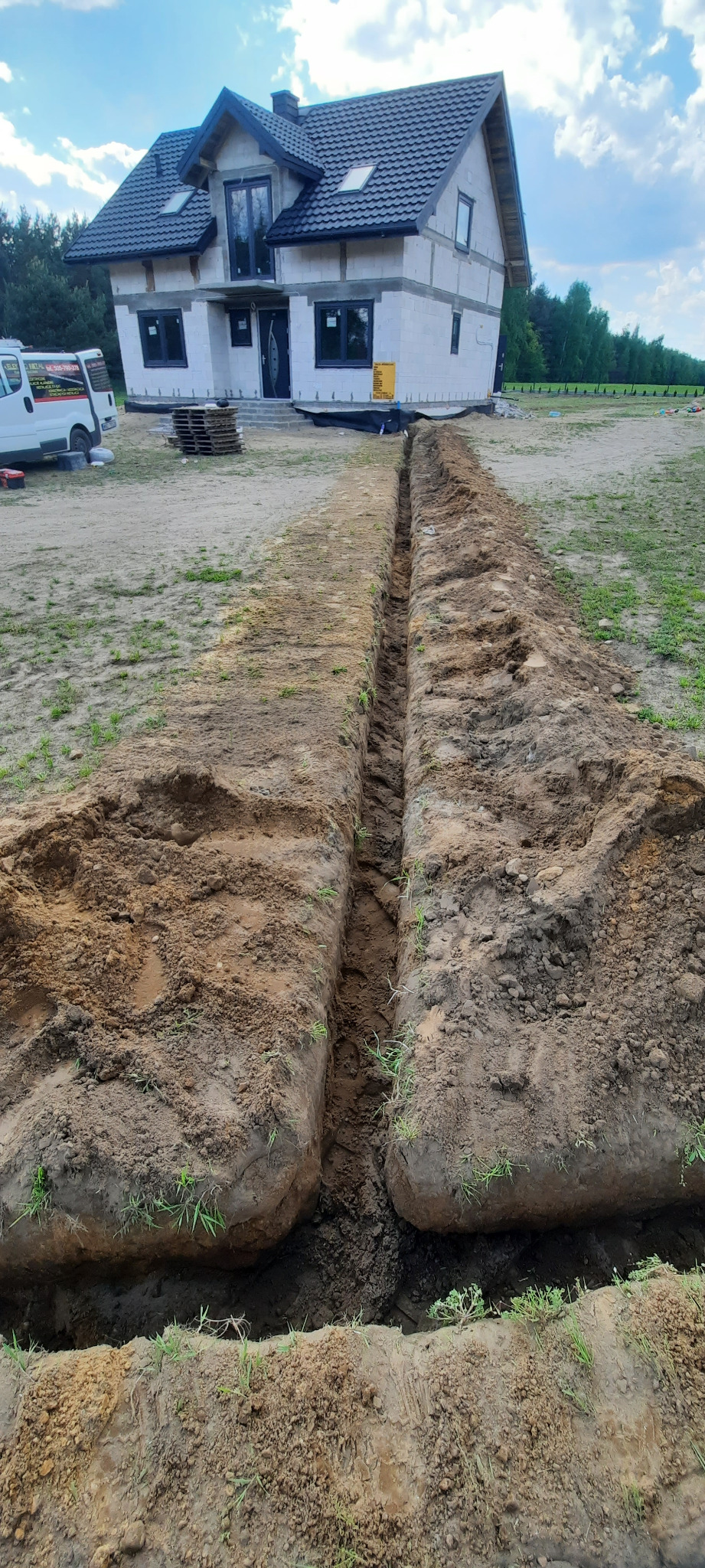 Wykopany rów w ziemi, prawdopodobnie pod kable elektryczne, prowadzący w stronę niedokończonego domu jednorodzinnego z białego pustaka, z widocznym samochodem dostawczym w tle.