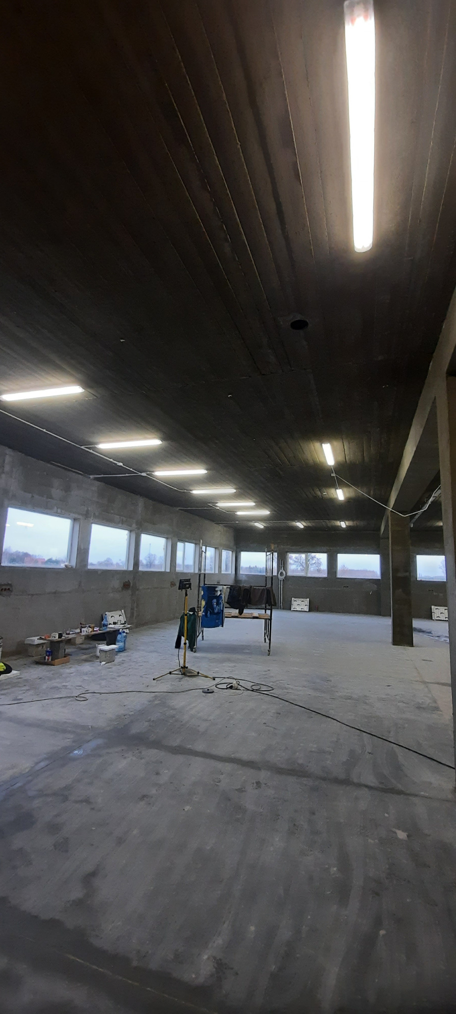 Wnętrze surowego, betonowego pomieszczenia z oświetleniem jarzeniowym; widoczne okna, narzędzia i prowizoryczne stanowisko pracy