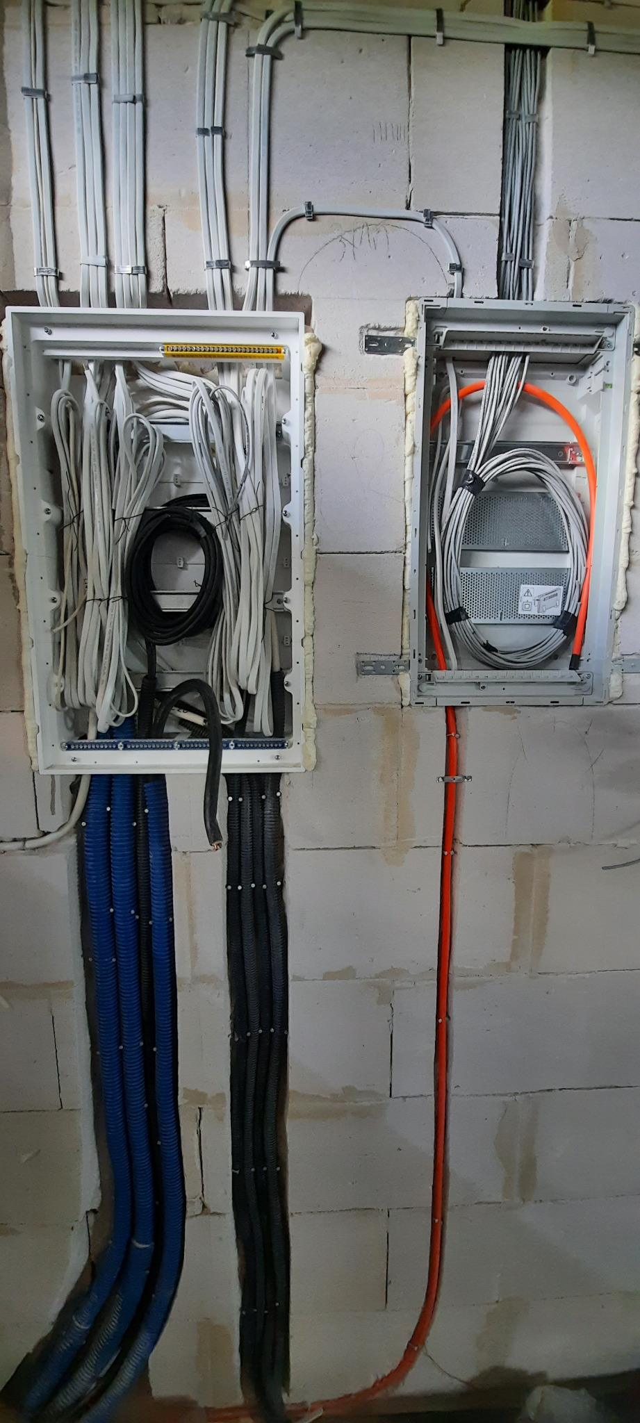 Dwie białe skrzynki elektryczne zamontowane na surowej ścianie z betonu komórkowego. Wewnątrz skrzynki widoczne liczne przewody elektryczne w różnych kolorach, w tym pomarańczowe rury karbowane.