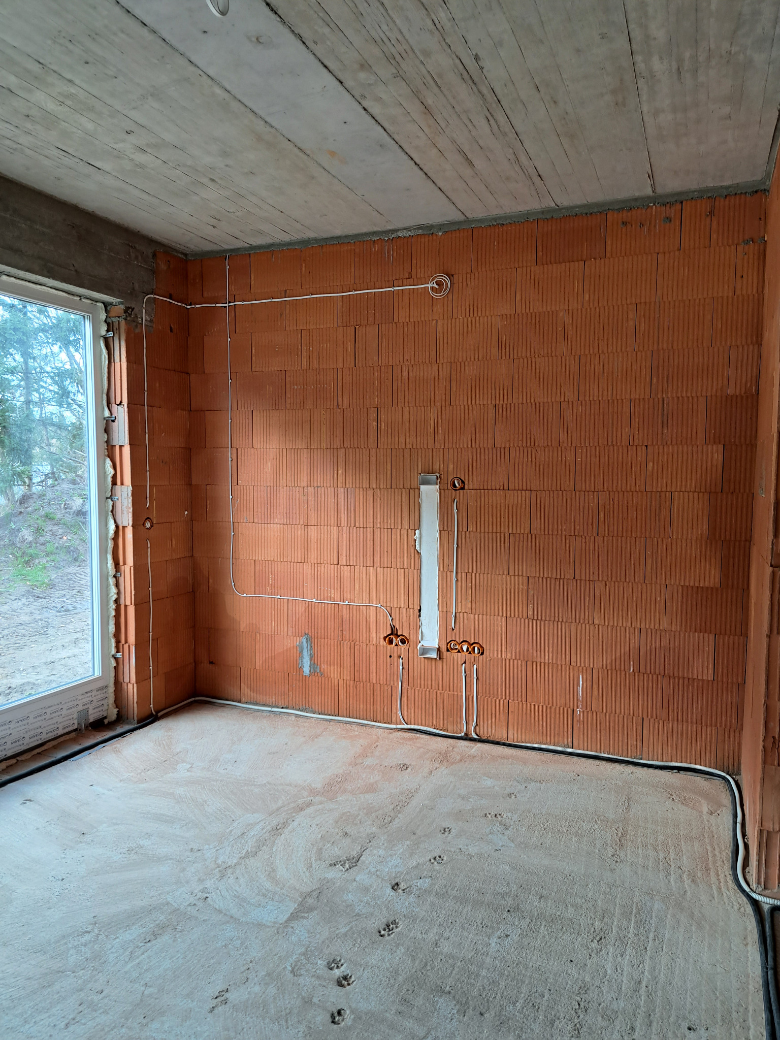 Surowy stan wnętrza z widocznymi przewodami elektrycznymi ułożonymi wzdłuż ścian z cegły, pod betonowym stropem; widoczne ślady łap na podłodze.