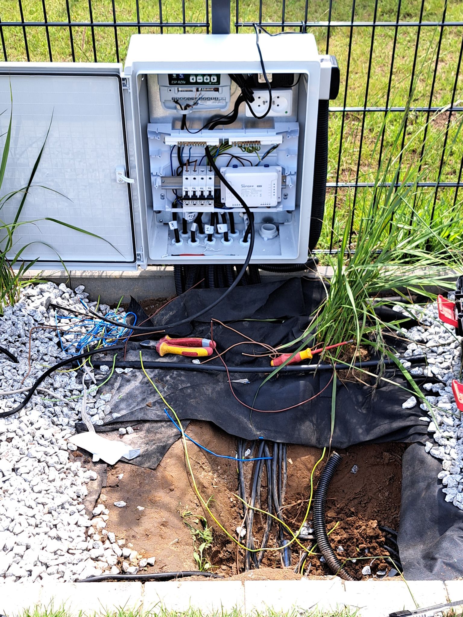 Otwarta skrzynka elektryczna z widocznym okablowaniem i elementami sterującymi, umieszczona w ziemi z kamieniami i fragmentem wykopu z rurami kablowymi.