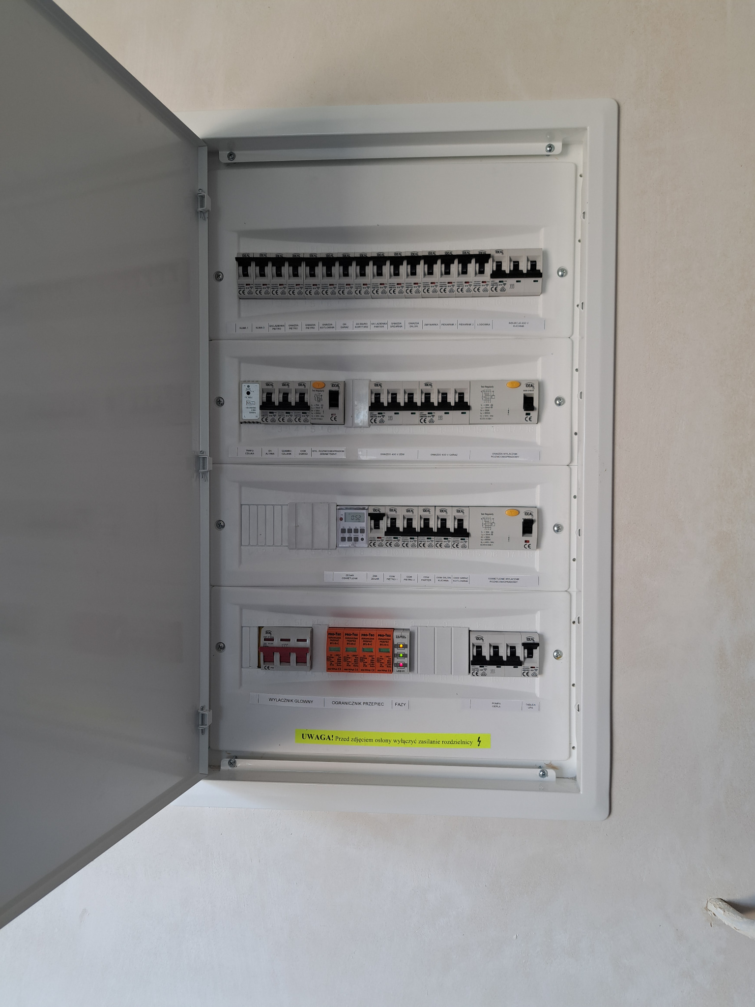 Otwarta skrzynka rozdzielcza z widocznymi bezpiecznikami, wyłącznikami różnicowoprądowymi i licznikiem energii elektrycznej. Na dole naklejka ostrzegawcza w kolorze żółtym.