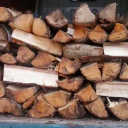 Stos pociętego drewna opałowego o nieregularnych kształtach, ułożonego warstwami w przestrzeni ładunkowej samochodu dostawczego.