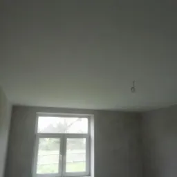 Pokój w trakcie remontu: świeżo wykończona, gładka, szara ściana i sufit, widoczne okno z widokiem na zewnątrz, wiszący kabel od lampy.