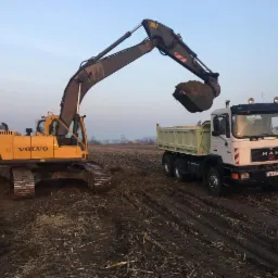 Żółta koparka Volvo nabiera ziemię do wywrotki MAN na polu, w tle widoczne drzewa i lekka mgła.