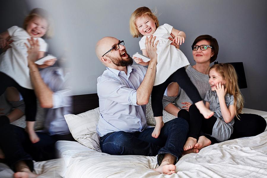 Radosna rodzina na łóżku: tata podrzuca uśmiechniętą córeczkę, mama siedzi z drugą córką, rozmyte odbicie taty i córki w tle.