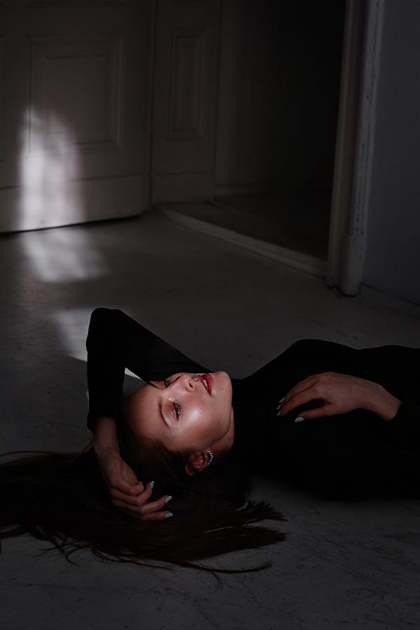 Kobieta w czarnym golfie leży na podłodze w słabo oświetlonym pomieszczeniu, światło pada na drzwi w tle, tworząc dramatyczny efekt.