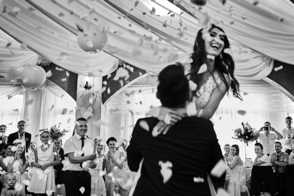Czarno-białe zdjęcie pary młodej w objęciach podczas pierwszego tańca, otoczonej gośćmi i opadającymi płatkami kwiatów, dekoracje sali z lampionami i białymi draperiami.