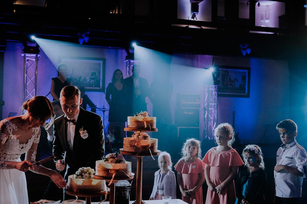 Para młoda kroi trzypiętrowy tort weselny na sali z podświetleniem, w tle dzieci i zespół muzyczny.