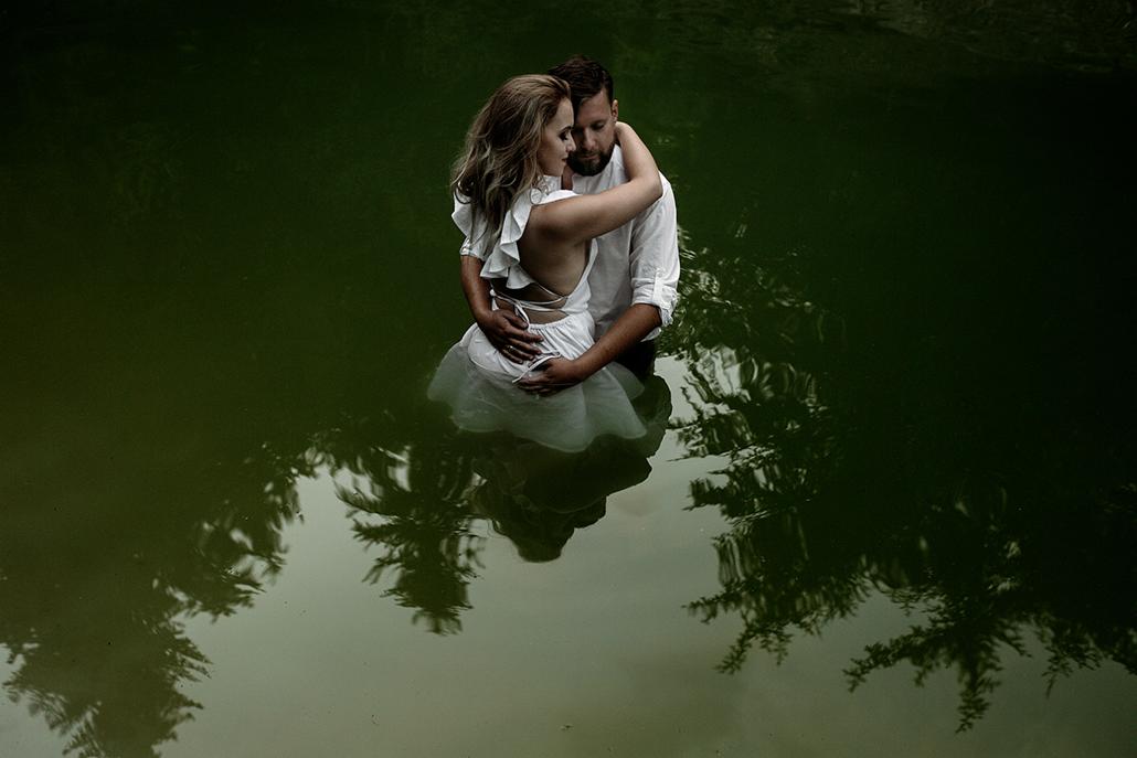 Para w białych ubraniach stoi w wodzie, odbicie drzew w ciemnozielonej powierzchni wody, romantyczna sesja plenerowa.