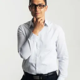 Portret uśmiechniętego mężczyzny w okularach, w jasnej koszuli i ciemnych spodniach, z ręką podpierającą brodę na jasnym tle.